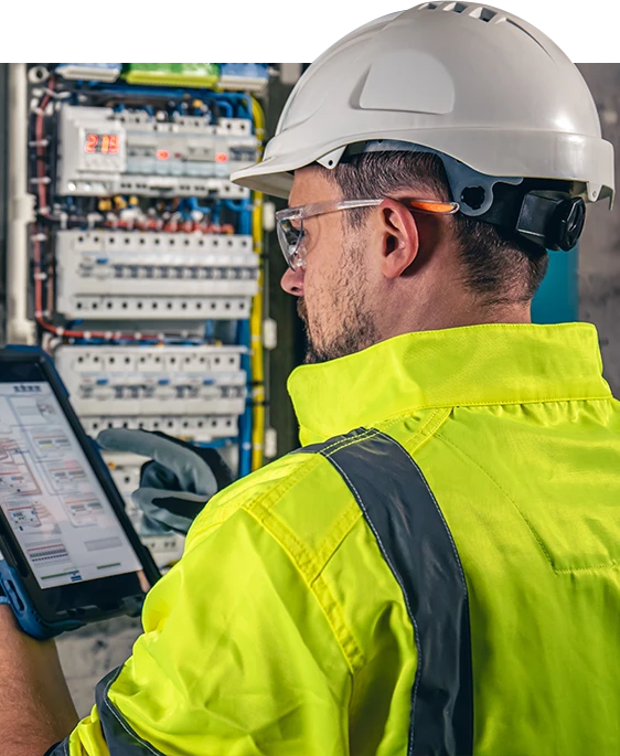 Engenheiro eletricista inspeciona painel elétrico utilizando tablet, usando EPI completo.